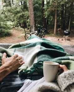 a person holding a coffee cup while laying on a blanket in the woods