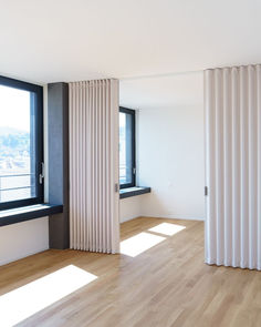 an empty room with wooden floors and white curtains on the windowsills, looking out onto a city