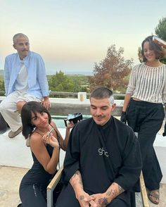 a man getting his hair cut while two women sit on the ground behind him and take pictures