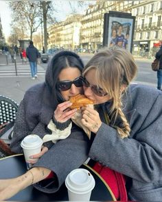 two women sitting at an outdoor table sharing a piece of bread and coffee in front of them