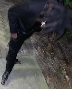 a man in black suit and tie standing on sidewalk next to tree with his foot up