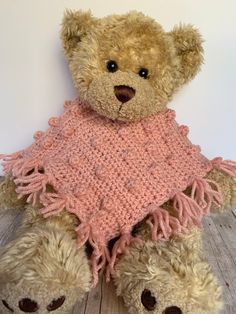 a brown teddy bear wearing a pink shawl on top of a wooden floor next to a white wall