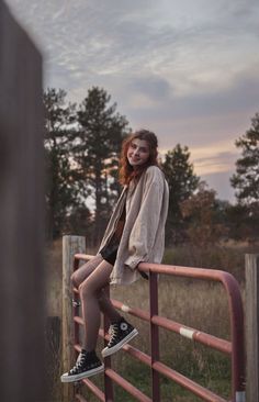 a young woman sitting on top of a metal rail next to a wooden fence with trees in the background