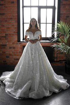 a woman standing in front of a window wearing a wedding dress with off the shoulder sleeves