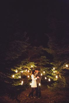 a man and woman standing in front of some trees with lights on them at night