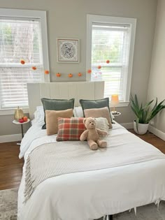 a teddy bear sitting on top of a white bed in a room with two windows