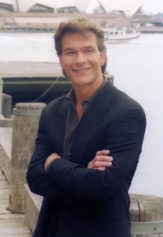 a man in a suit standing on a dock next to the water with his arms crossed