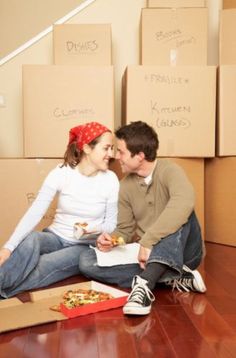 a young man and woman sitting on the floor eating pizza in front of moving boxes