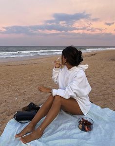 a woman sitting on top of a beach next to the ocean eating food and drinking