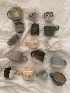 there are many cups and mugs on the white bed sheet, all in different shapes and sizes