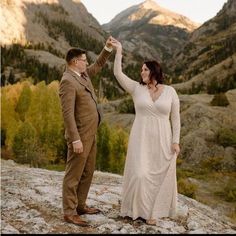 a man and woman holding hands while standing on top of a mountain with mountains in the background