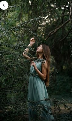 a woman standing in the woods with her eyes closed and hands on her head, looking up into the sky