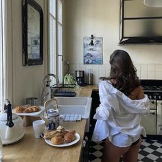 a woman standing in front of a kitchen counter with pastries on plates and coffee mugs