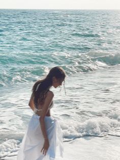 a woman in a white dress is standing on the beach and looking at the water