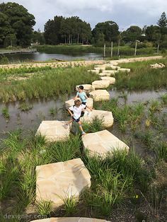 two children are sitting on stepping stones in the water near some grass and rocks that look like stepping stones