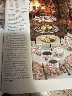 an open book with plates and cups on the table in front of a fire place