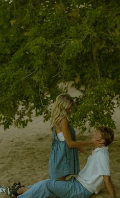 two people are sitting on the sand under a tree and one is touching the other's hand