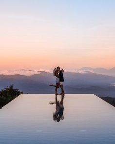 a man and woman kissing on the edge of a pool at sunset with mountains in the background