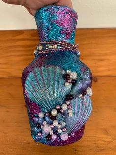 a hand holding a purple and blue vase with pearls on it's sides, sitting on a wooden table