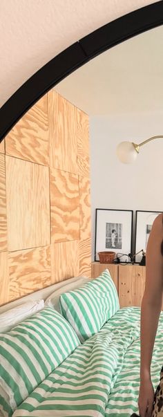 a woman standing in front of a mirror on top of a wooden headboard next to a green and white striped bedspread