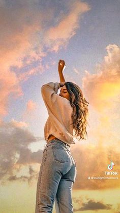 a woman standing on top of a hill with her arms in the air and clouds behind her