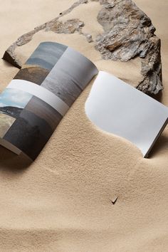 an open book sitting on top of a sandy beach next to a rock and paper