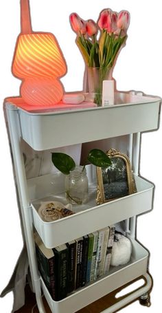 a white shelf with some flowers and books on it's sides, next to a lamp