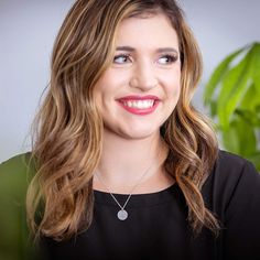 a woman with long brown hair wearing a black top and smiling at the camera while standing in front of a potted plant