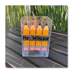 a clear box with colored pencils in it on top of a wooden table next to grass