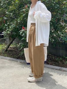 a woman standing in front of a bush with her hands on her hips, wearing brown pants and a white shirt