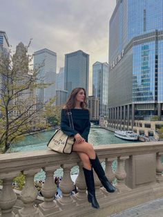 a woman is sitting on a railing in front of the water and skyscrapers with her legs crossed