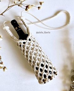 a white crocheted bag with a black bottle in it next to some flowers