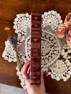 a person holding a book on top of a doily with flowers in the background