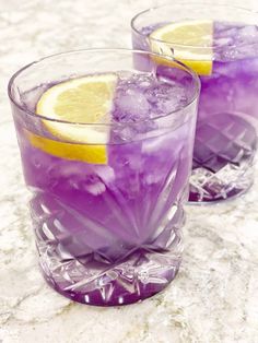 two glasses filled with purple liquid and lemon wedges on top of a marble table