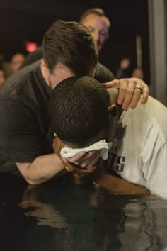 two men hugging each other in front of a crowd at a bar or restaurant, with one man covering his face and the other leaning over