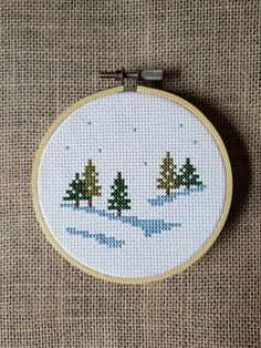 a cross stitch christmas scene with trees in the snow on a beige background, hanging from a wooden hoop