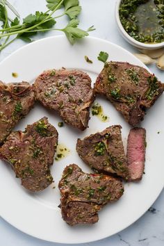 some meat is on a white plate next to green herbs and seasoning sprinkled with parsley