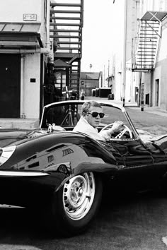 a black and white photo of a man driving a sports car down the street in front of a tall building