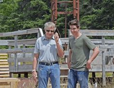 two men standing next to each other in front of a wooden structure with trees behind them