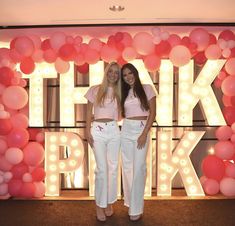 two young women standing in front of a giant sign with balloons on the wall behind them