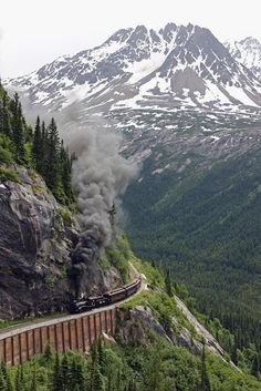 a train traveling down tracks next to a mountain covered in trees and snow on top of it