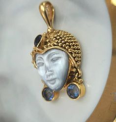 Sajen is the Balinese word for "offerings," and the trademark name for Sajen jewelry creations. This Balinese Goddess  wears an ornate gold beaded headdress with  a bezeld Iolite pear shape gemstone  over her forehead. She also wears opulent round blue sapphire dangling earrings. This is one piece of mother of pearl carving interictally detailed. 11.0 grams of 18K yellow gold The bale accommodates most chains 4.7mm width of space for a chain. This item comes wrapped and shipped in a Fedex Package including insurance Exquisite Carved Gold Jewelry, Gold Tanzanite Pendant Jewelry, Gold Tanzanite Jewelry For Wedding, Jewelry Creation, Gold Beads, Mother Of Pearl, Blue Sapphire, Bali, Sapphire