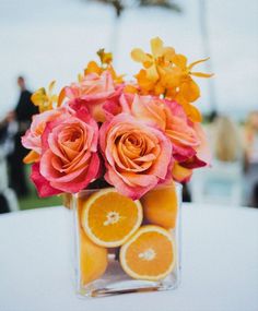 an arrangement of oranges and roses in a square vase on a table with people