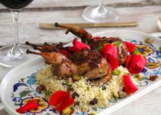 two pieces of meat on top of rice with red flowers and wine glasses in the background