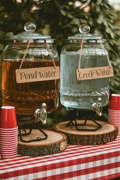 One Happy Camper 1st birthday party drink station featuring glass dispensers labeled 'Pond Water' and 'Creek Water,' set on rustic wood slices. The setup includes a red and white checkered tablecloth, perfect for an outdoorsy camping them first birthday celebration. 1 Happy Camper Birthday, Birthday Creative Ideas, One Happy Camper First Birthday, Camping Baby Shower Theme