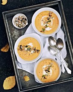 three bowls of soup on a tray with silver spoons and autumn leaves around them