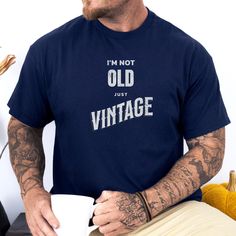 Celebrate your timeless style with this fun and bold Gildan 5000 t-shirt! Featuring the playful phrase, "I'm Not Old, Just Vintage," this shirt is perfect for those who embrace their age with confidence and a touch of humor. Whether you're gifting it to someone or adding it to your own wardrobe, this tee makes a statement in style! Product Features: Graphic: "I'm Not Old, Just Vintage" - a quirky design that stands out with its retro-inspired typography. Material: Made from 100% cotton for a sof Vintage Birthday T-shirt With Text Print, Classic Graphic Print T-shirt For Birthday, Vintage Cotton T-shirt With Funny Text, Vintage Cotton Tops For Anniversary, Vintage Fan Merchandise T-shirt With Letter Print, Retro Short Sleeve T-shirt For Birthday, Anniversary Tops With Funny Text And Short Sleeves, Vintage Text Print Top For Birthday, Vintage Slogan T-shirt For Fans