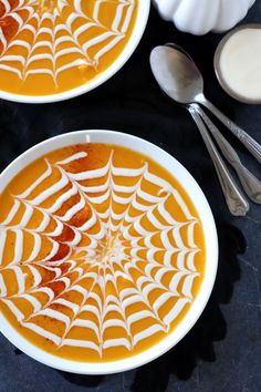 two white bowls filled with pumpkin soup on top of a black table next to silver spoons