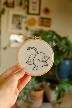 a hand holding up a small embroidered bird on a wooden frame with potted plants in the background