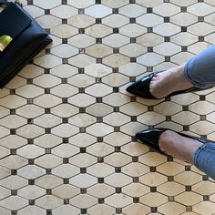 a woman's feet in black shoes standing on a tiled floor next to a purse